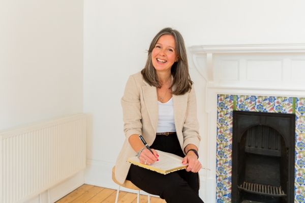 Gemma Pettman, a white woman with shoulder length dark blonde hair sits on a chair in a white room with a tiled fireplace in the background. She is wearing black trousers, a white top and cream jacket and has a notepad on her lap.